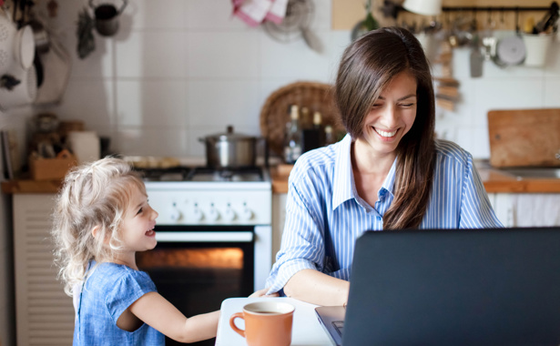 A woman sits at a laptop in her kitchen accessing the internet via her home broadband as her child watches on